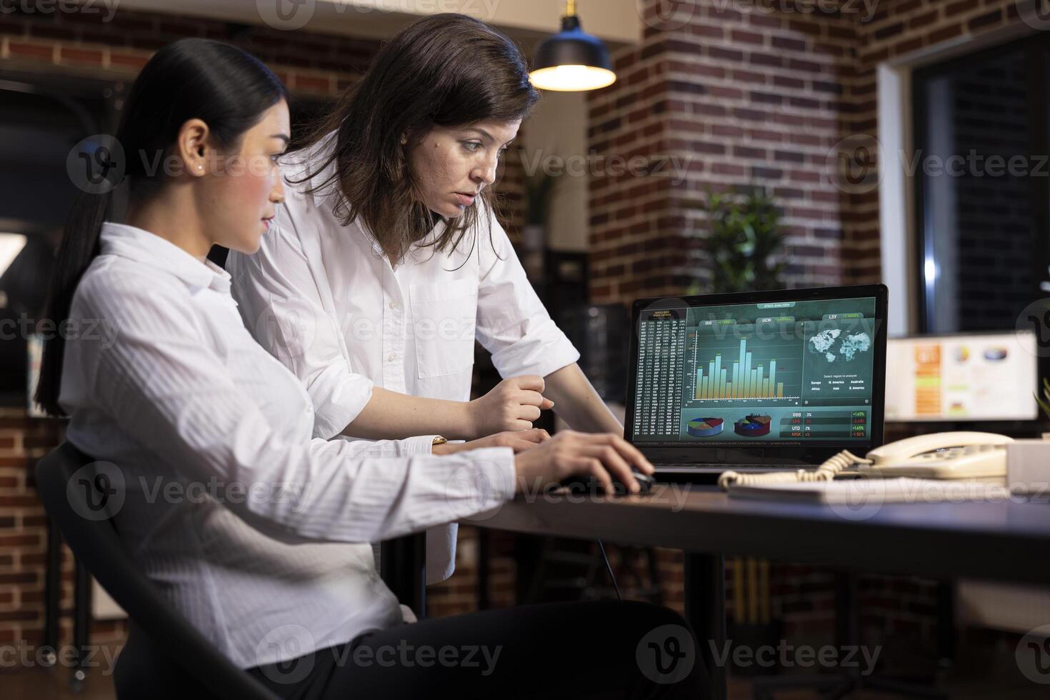 A professional discussion between coworkers, collaborating on a business project, using research data on laptop. White woman stands and reviews details as her colleagues scrolls through the document. photo