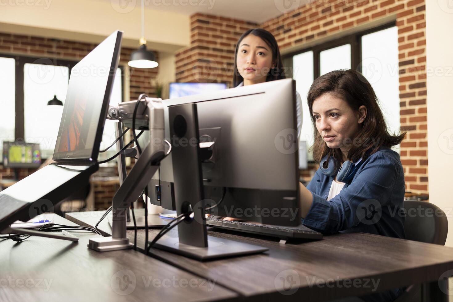 Selective focus on caucasian woman focused on computer screen, reviewing product designs in brick wall office. Female graphics designer analyzing images on desktop pc as asian manager stands nearby. photo