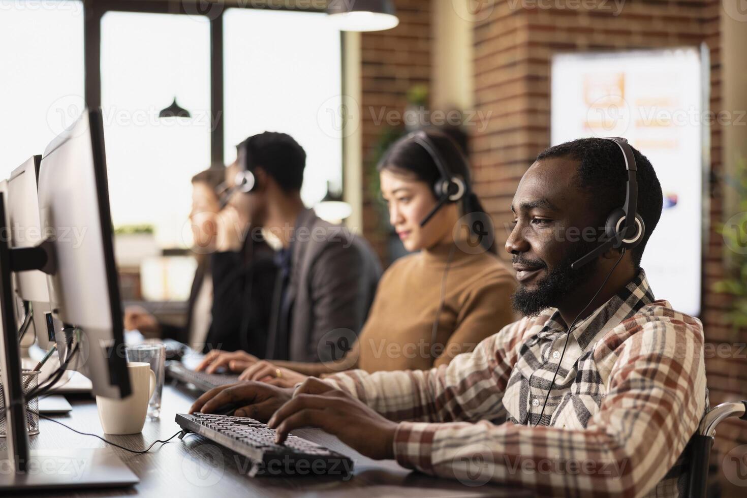 Black man in wheelchair and his multiracial coworkers seated at desks with computers and headsets, handling client calls. African American male customer service agent working in brick wall workspace. photo