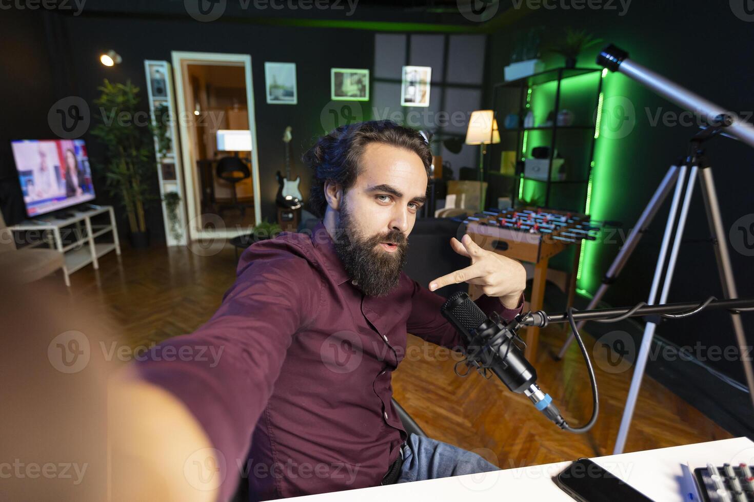 Influencer in home studio using smartphone on selfie stick to record live broadcast for online streaming platforms, talking with fans. Smiling media star filing himself doing cool hand gesturing photo