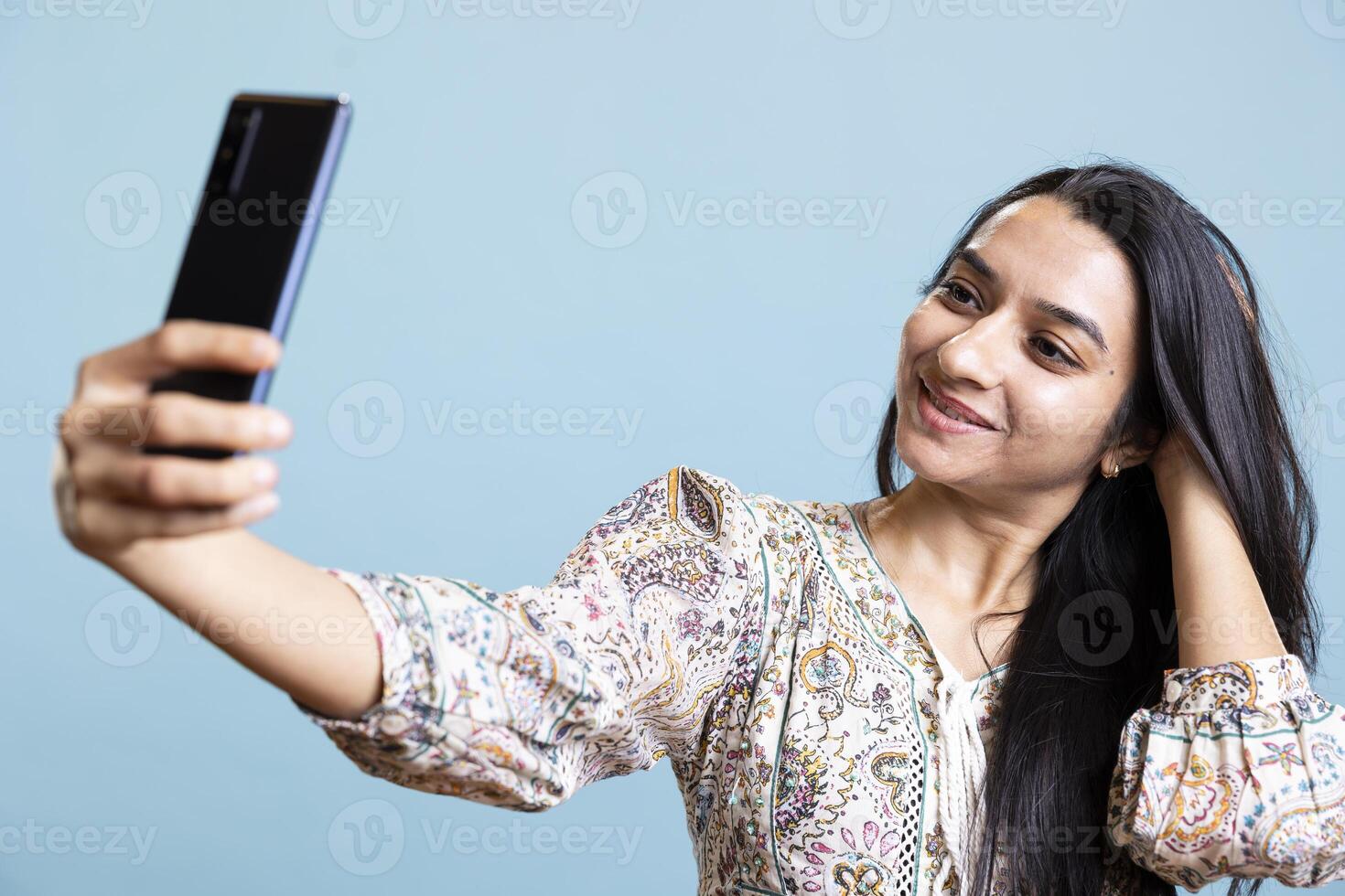 Pleased cheerful person using her smartphone to take selfies on social media. Jolly indian woman person taking photos using phone camera, standing against studio blue background.