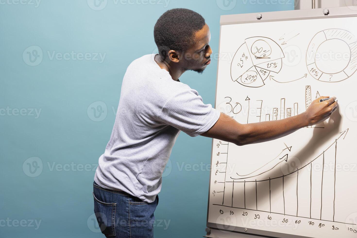 African american male professional in casual clothing drawing graphs and charts on a sketch board. Black man standing next to whiteboard and preparing data for presentation in blue background studio. photo