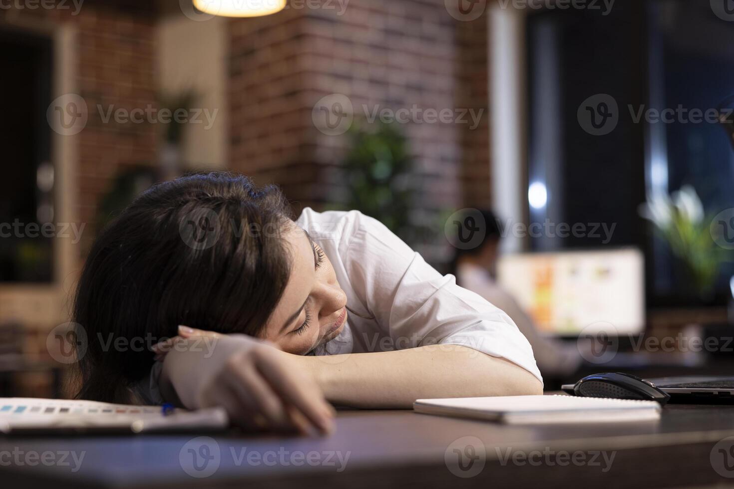 Tired businesswoman takes powernap at her workstation, resting briefly amid her demanding schedule in late night office setting. Caucasian entrepreneur dozes off at desk, exhausted from her workload. photo