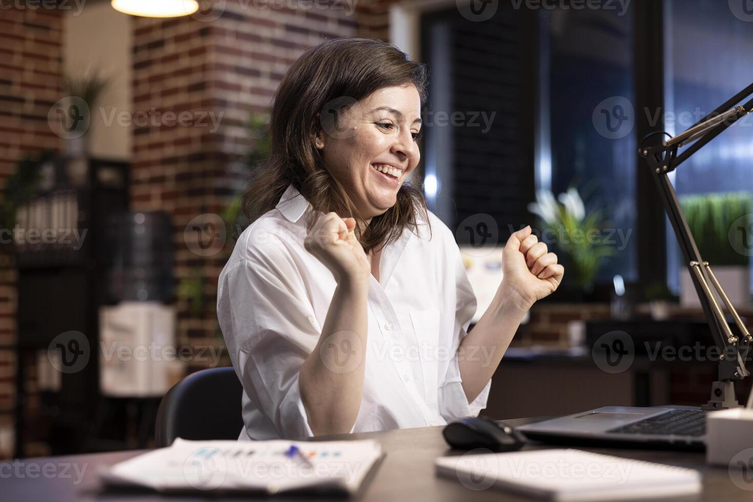 Professional woman celebrates career milestone at desk, dancing in her chair with arms raised, smiling as she looks at her laptop. Excited female entrepreneur feeling triumphant after an achievement. photo