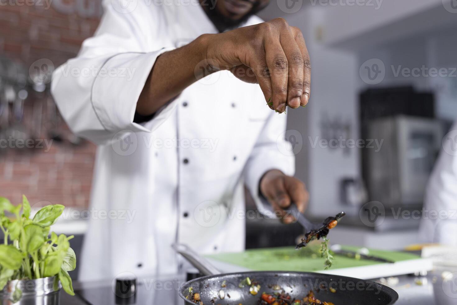 Black head chef adding a sprinkle of rosemary to a gourmet dish on the stove, using fresh produce and herbs to enhance the meals flavors. Culinary arts and gastronomy, chef life. photo