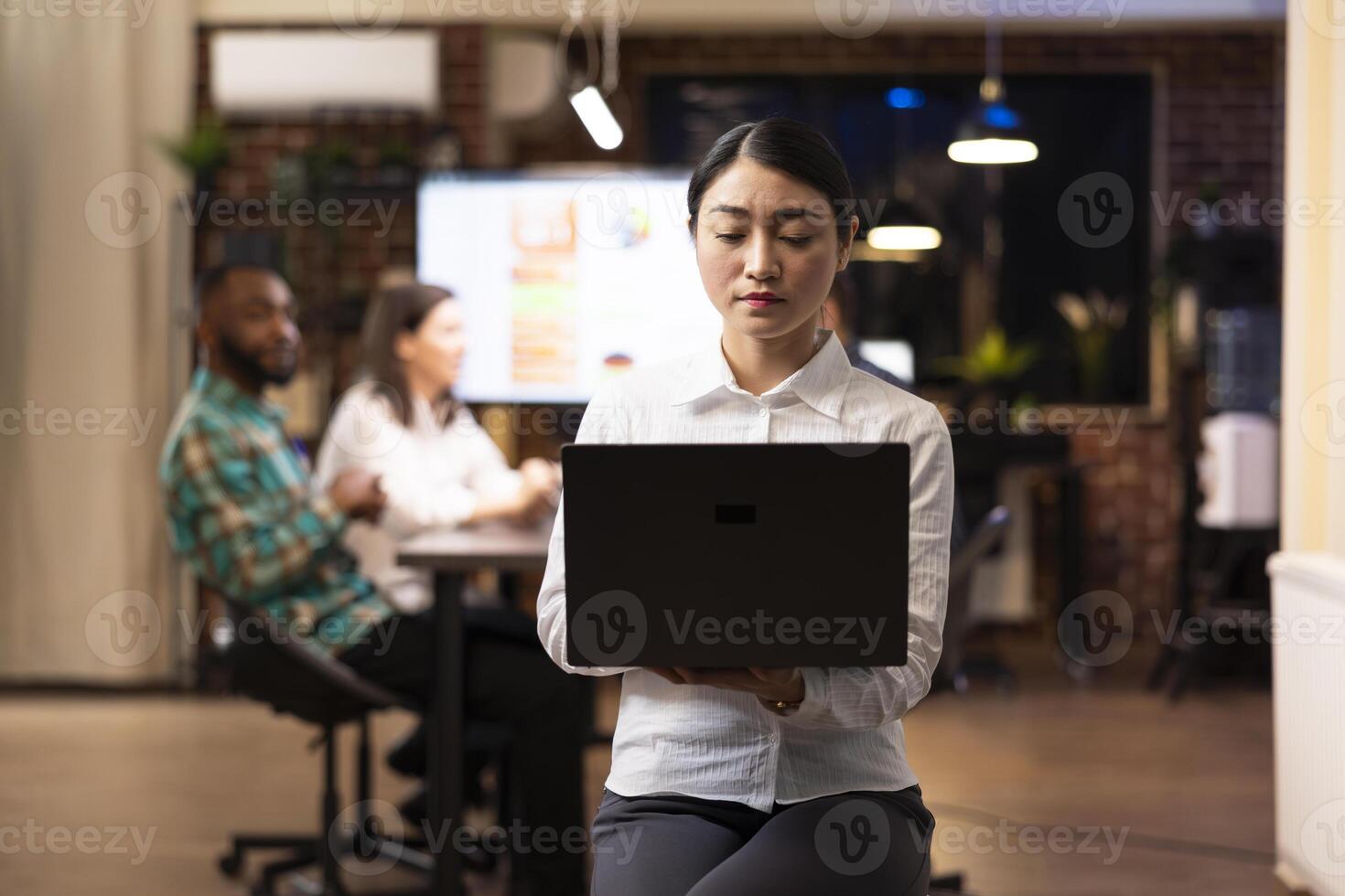 Focused asian businesswoman working on laptop, analyzing marketing trends as her coworkers discuss financial charts in background. The late night office setting highlights productivity. photo