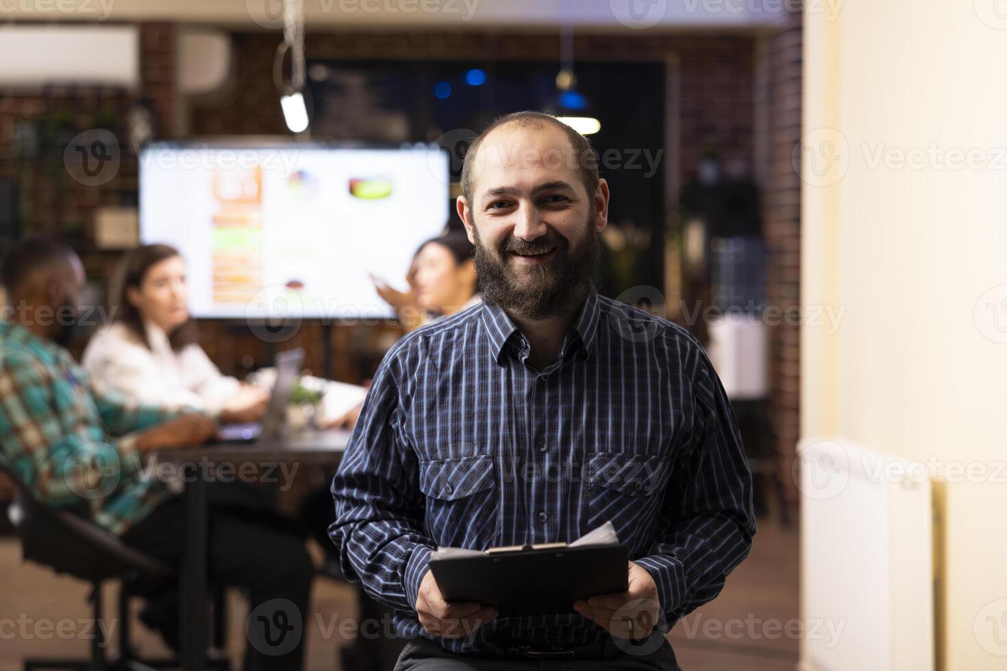 Caucasian male entrepreneur holds a clipboard in modern startup office. The night shift setting, brick wall and blurred team in background emphasize productivity and teamwork. photo