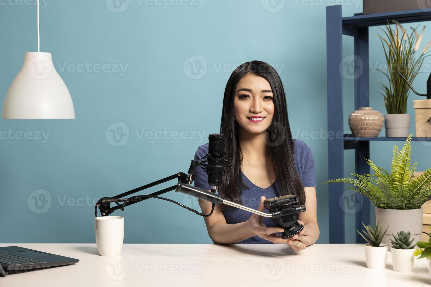 Asian female vlogger presenting a camera tripod as she speaks into a microphone in a professional studio setup. Young woman seated at her desk, reviewing filming equipment for content creation. photo