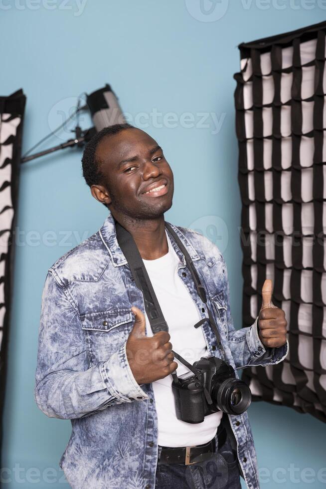 Happy male photographer showing two thumbs up after successful client photoshoot session in studio. African american man in casual clothing and dslr camera on his neck, reflecting positivity. photo