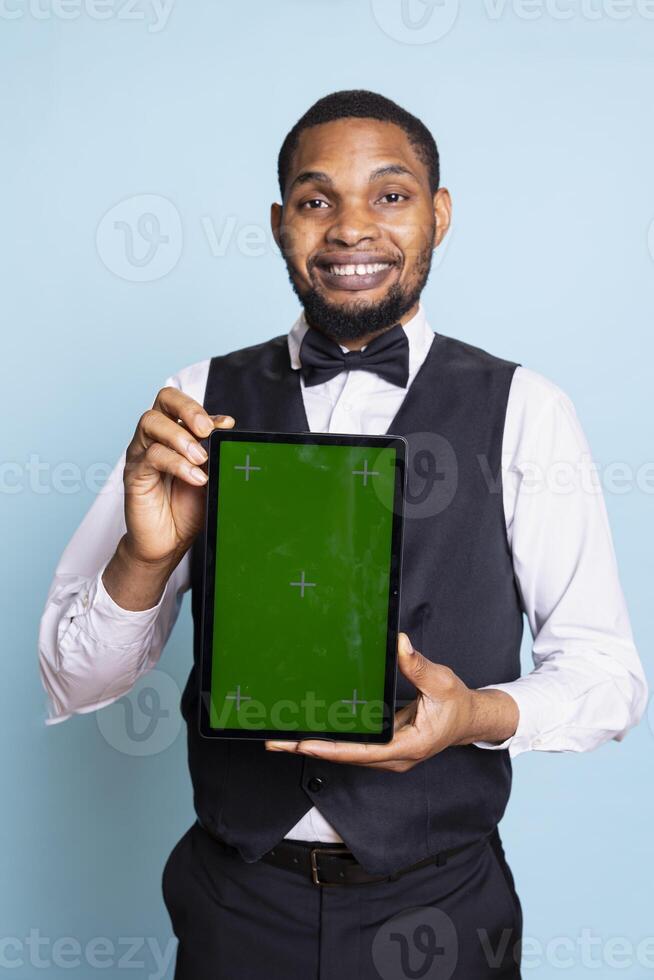 Smiling hotelier holding a tablet with isolated chroma key screen in studio, wearing a professional tuxedo uniform and greeting clients. Young hotel concierge against blue background. photo