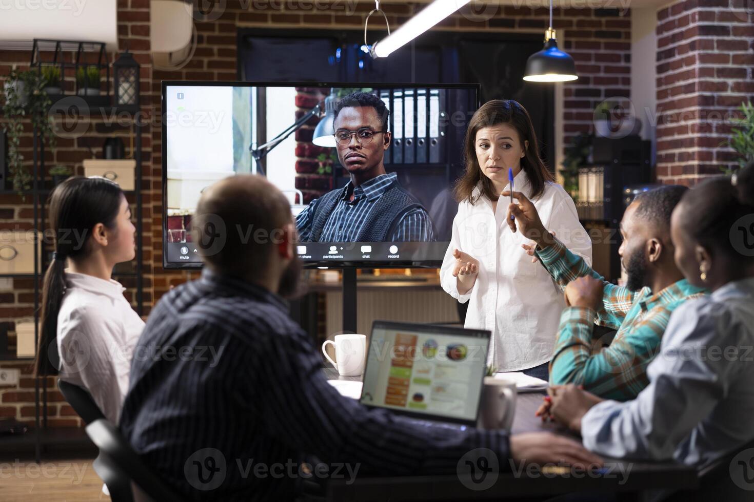 Multiethnic startup team in modern office with brick wall background, engaged in late night business meeting. A digital screen displays call with remote african american manager. photo
