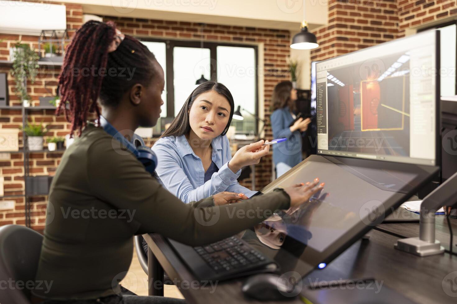 Black female employee sits at desk, updating her asian creative manager on latest progress of the project. Professional women editors collaborate on startup design, sharing ideas in brick wall office. photo