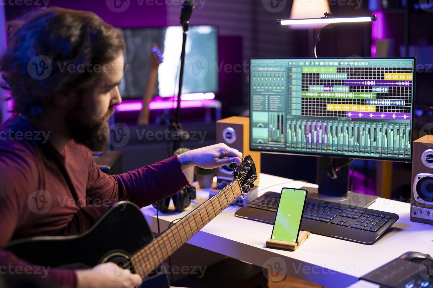 Sound engineer learns tuning acoustic guitar in home studio, watching a tutorial on internet via mobile phone with greenscreen. Artist learning to play instrument and create tunes. photo