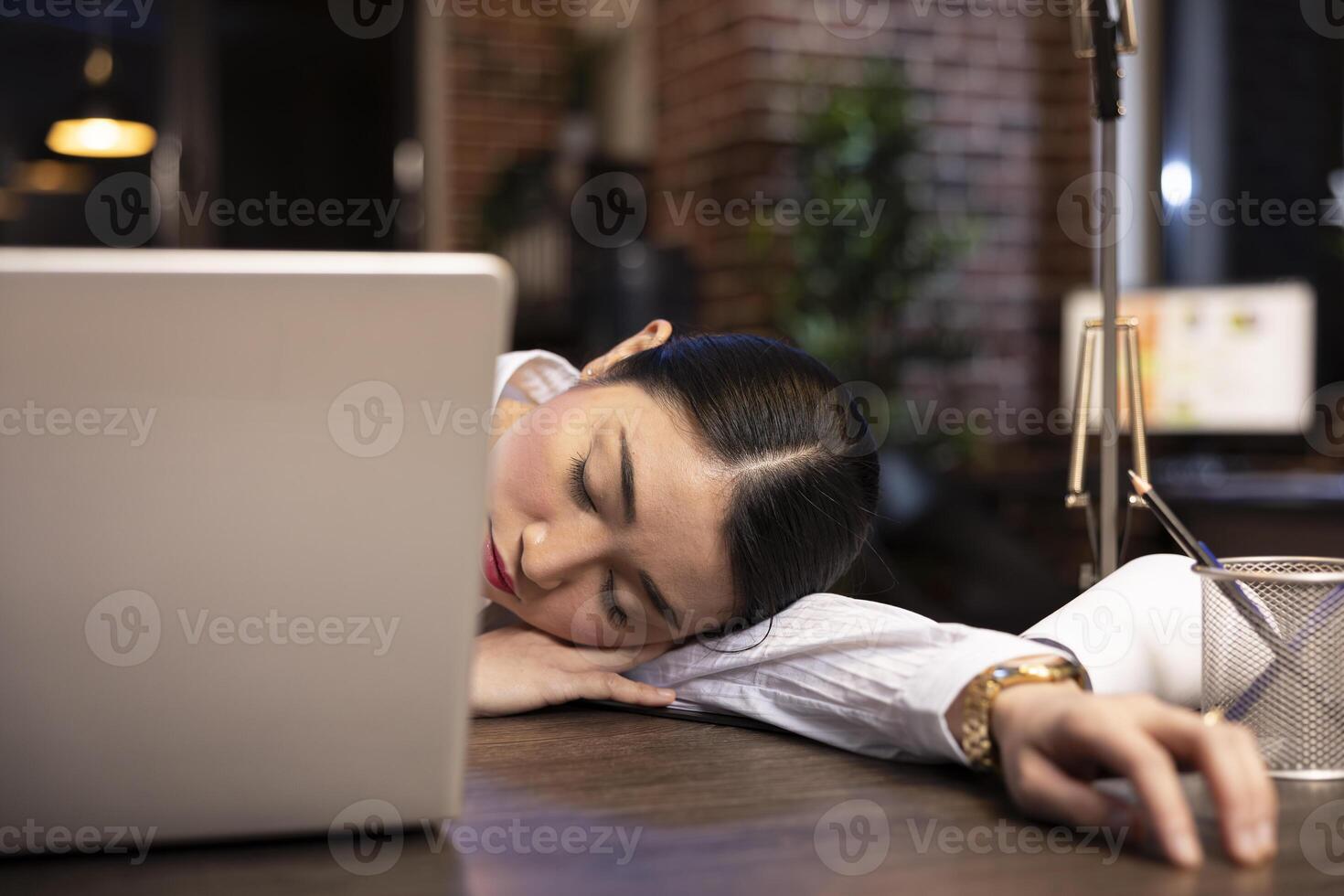 Asian female entrepreneur falls asleep at her desk during a late night shift in brick wall office. Tired businesswoman takes a brief rest amidst her busy schedule as her laptop remains open on table. photo