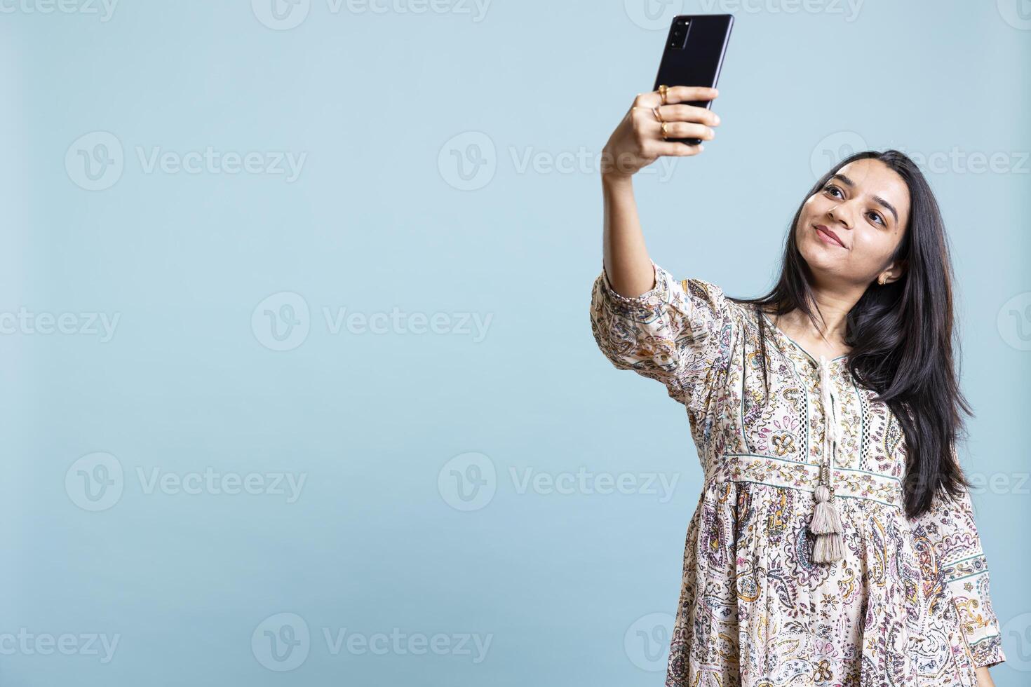 Jolly indian woman smiling for selfies posting them on social media profile, feeling confident and pretty in the studio. Happy silly girl taking pictures using phone selfie camera, blue background. photo