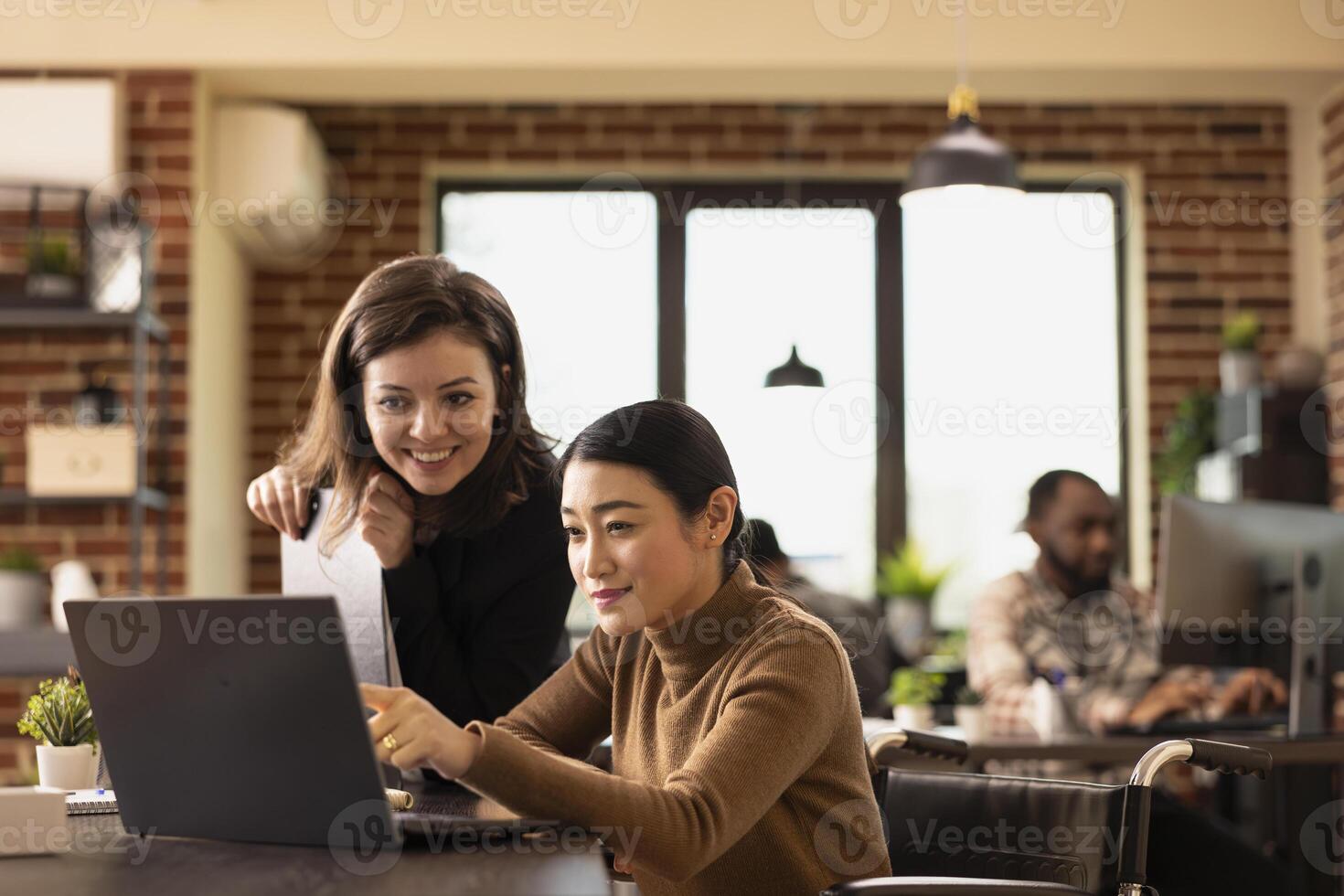 Multiethnic employees working together at office desk, reviewing financial data on laptop. An inclusive workspace where caucasian manager and asian wheelchair user discuss business growth strategies. photo