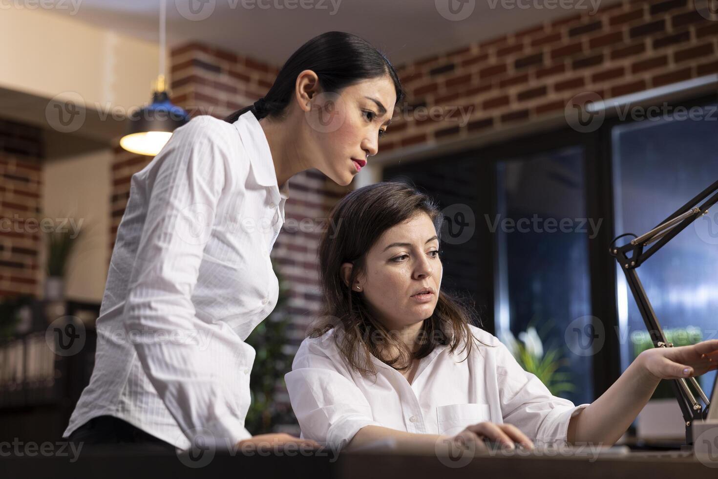 Dedicated female employees analyze research data on a laptop, having a business discussion. Asian manager stands and listens as her coworker explains the details, working together on a business task. photo