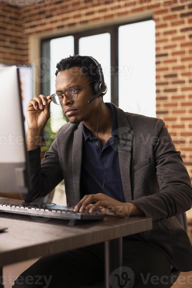 Pensive black man adjusting his glasses and working at desktop pc, focused on client information. Concentrated call center agent wearing headset, attending to customer support tasks on his computer. photo