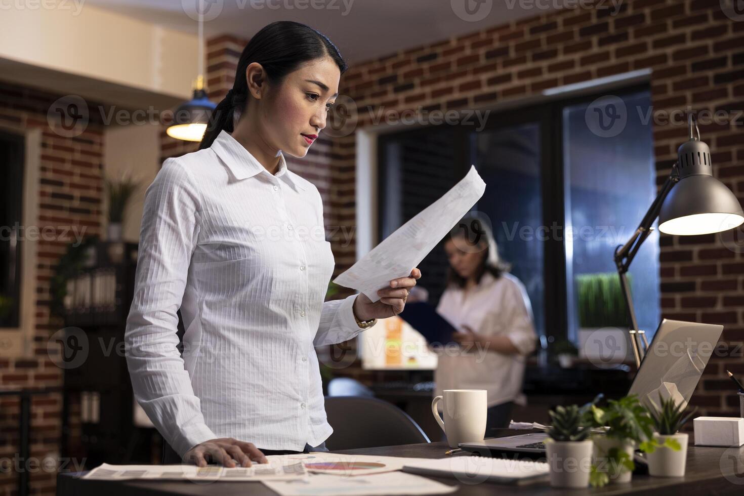 Focused employee reading a document in hand, standing at her office desk during her night shift. Asian corporate manager on her feet, working late, planning a brand strategy for startup company. photo