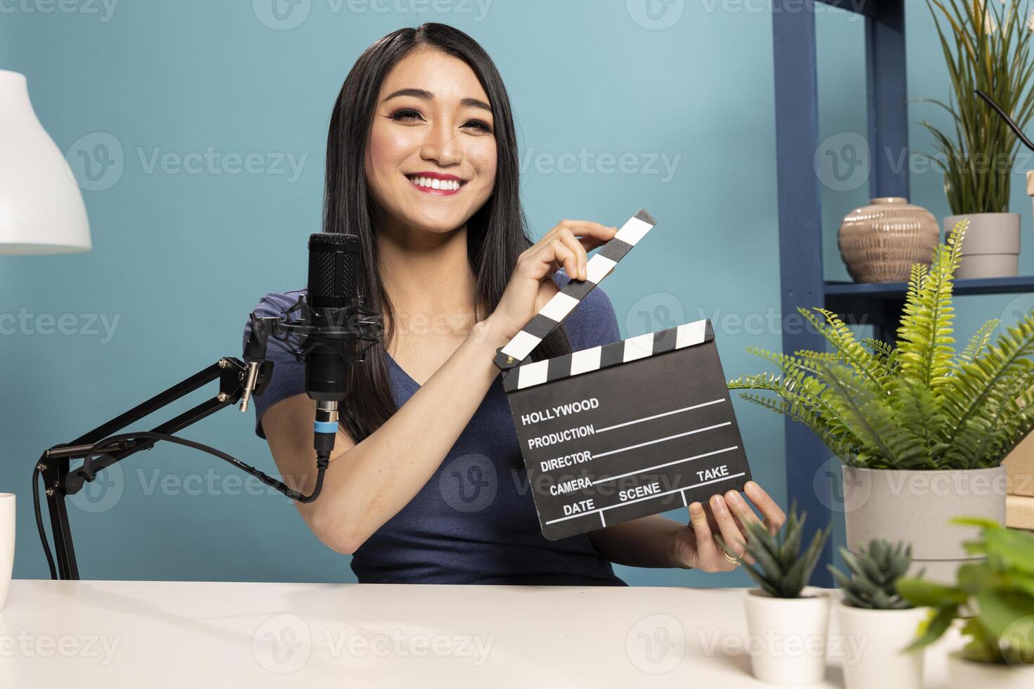 Female vlogger presents a clapperboard into the camera, sitting at a desk with a microphone. The scene highlights filmmaking equipment and atmosphere of content creation for online reviews. photo