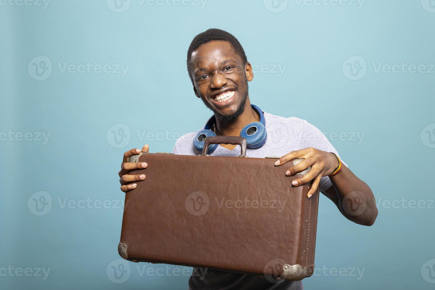 Cheerful african american man with a suitcase and headphones, excited for his journey. Black male tourist looking at the camera and posing with his luggage against an isolated background. photo
