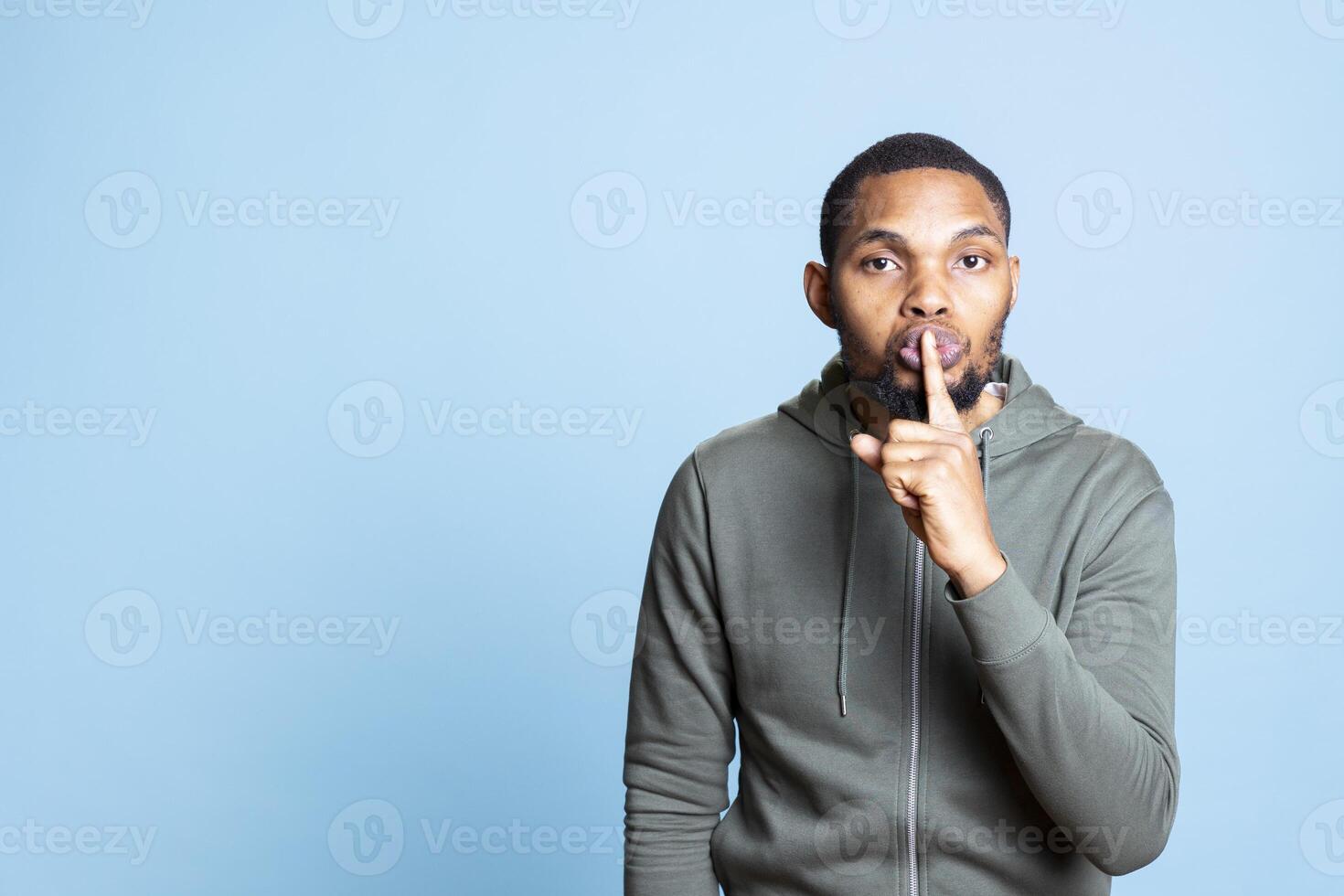African american confidential man asking for silence and secrecy against blue background, keeping a secret and asking for privacy. Young silent person showing hush symbol with finger over lips. photo