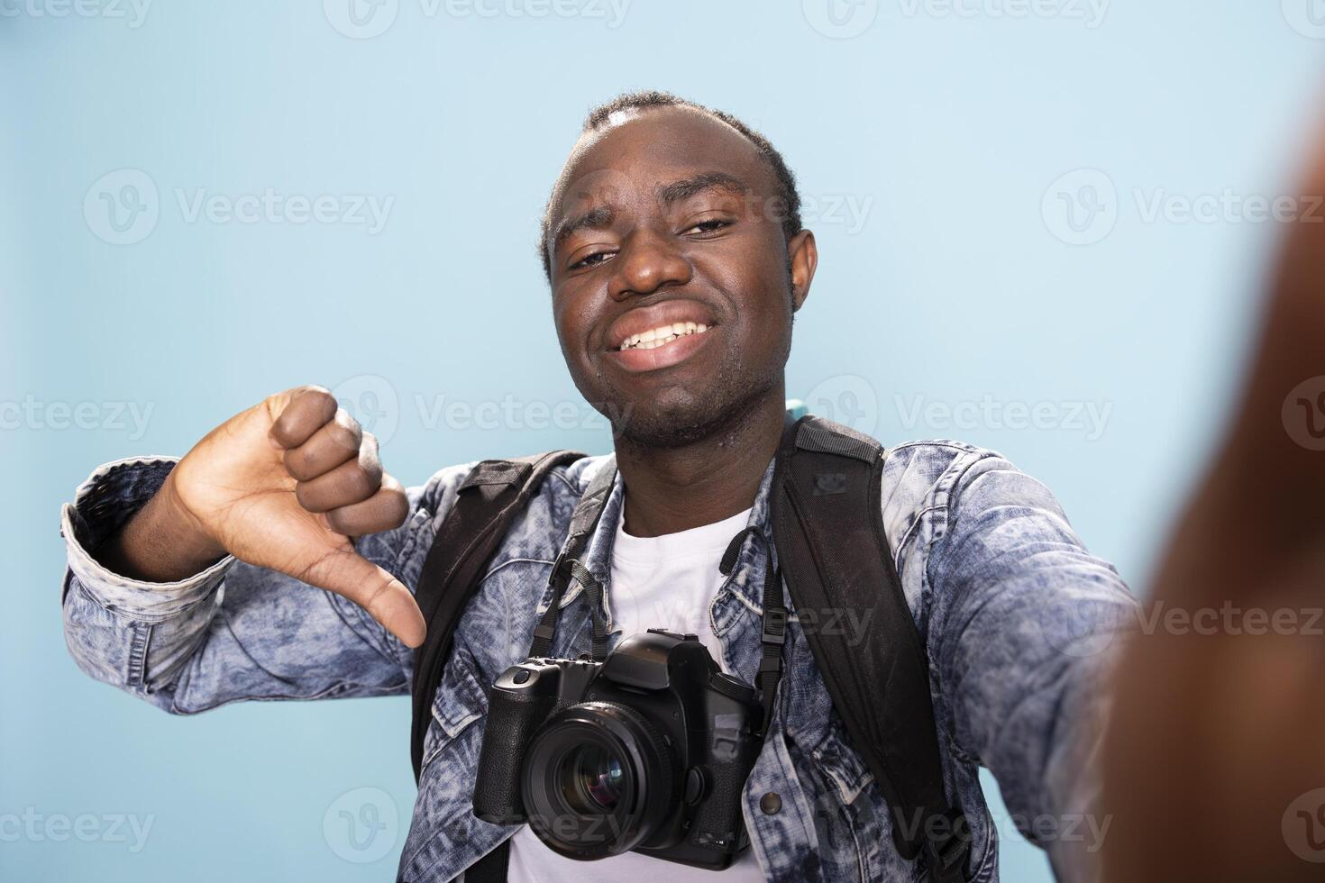 Happy african american photographer with a professional camera around his neck, giving thumbs down in studio. POV of a black male vlogger recording himself while making a playful disagreeing gesture. photo