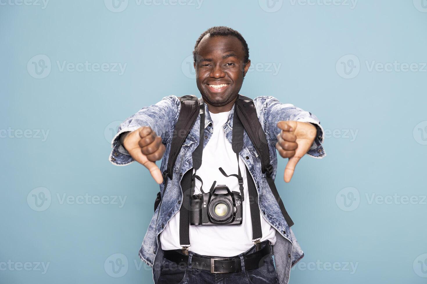 African American man in casual clothing, cheerfully giving two thumbs down while standing against isolated background. Smiling black male photographer with dslr camera, expresses disapproval in studio. photo