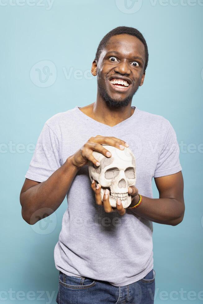 African american man with wide eyes, holding a skull and making goofy expressions as he looks at camera. Young black male individual grasping a human skeletal head against an isolated blue background. photo