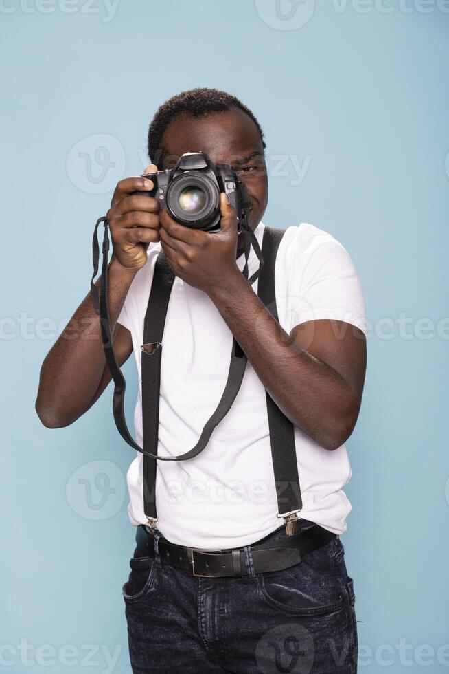Black photographer wears suspenders, standing in a blue background studio and pointing his camera forward, ready to capture the perfect shot. His determined expression highlights his creative vision. photo