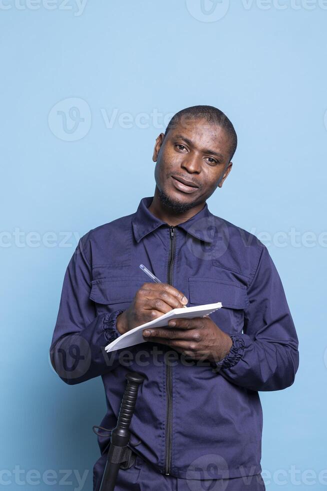 Security agent in safeguarding uniform takes notes on a notebook, standing against blue background. Serious dedicated bodyguard writing info to keep track of safety responsibilities. photo