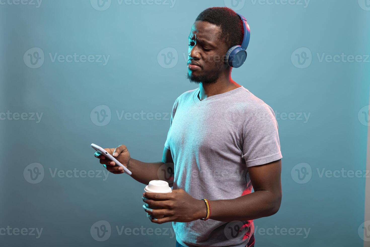 Young male individual holding a disposable cup and looking at his cellphone with a confused expression. Black man with wireless headphones unsure with a on his mobile device. photo