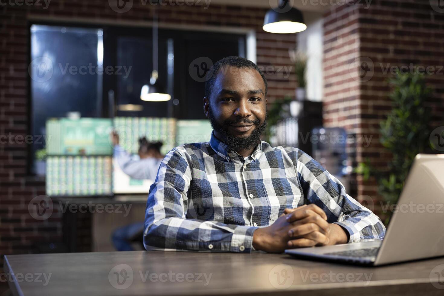 Portrait of african american man smiling and looking at the camera, seated at a desk with a laptop. The office is dimly lit with a brick wall, ideal for night shift work and stock market monitoring. photo