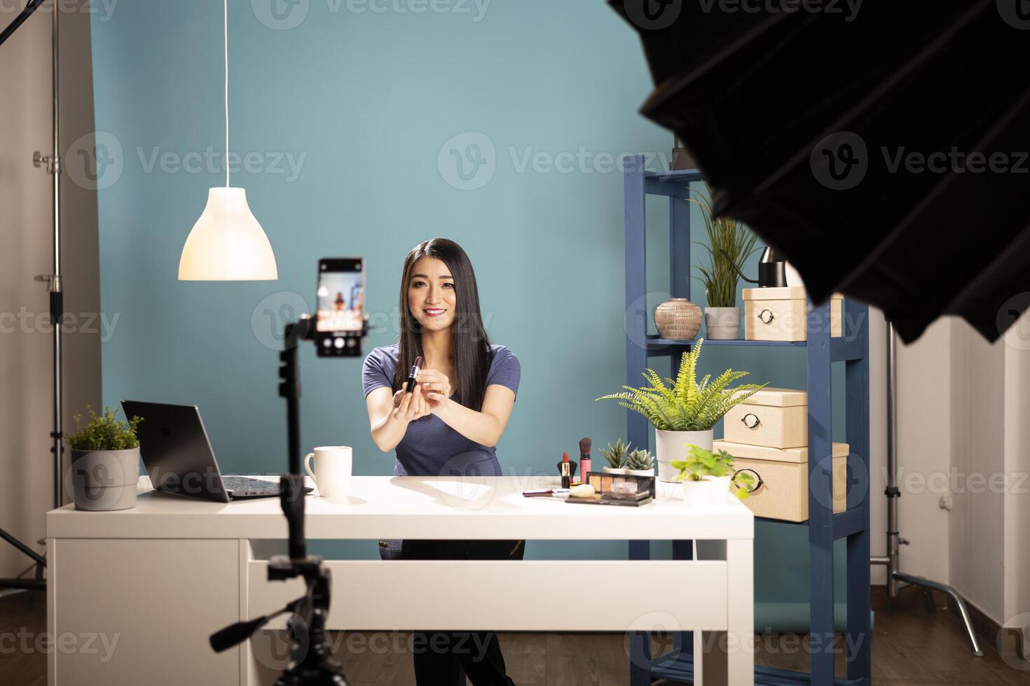Professional beauty influencer demonstrating skincare and makeup products for online tutorial. Asian female vlogger with lipstick in hand is capturing high quality content using smartphone and tripod. photo