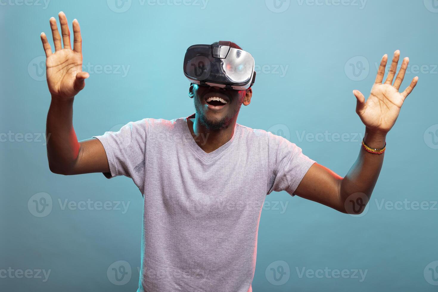 Millennial man raising hands and playing a futuristic game, enjoying immersive 3D simulation. African american person with an amazed expression, using VR glasses against a blue background. photo