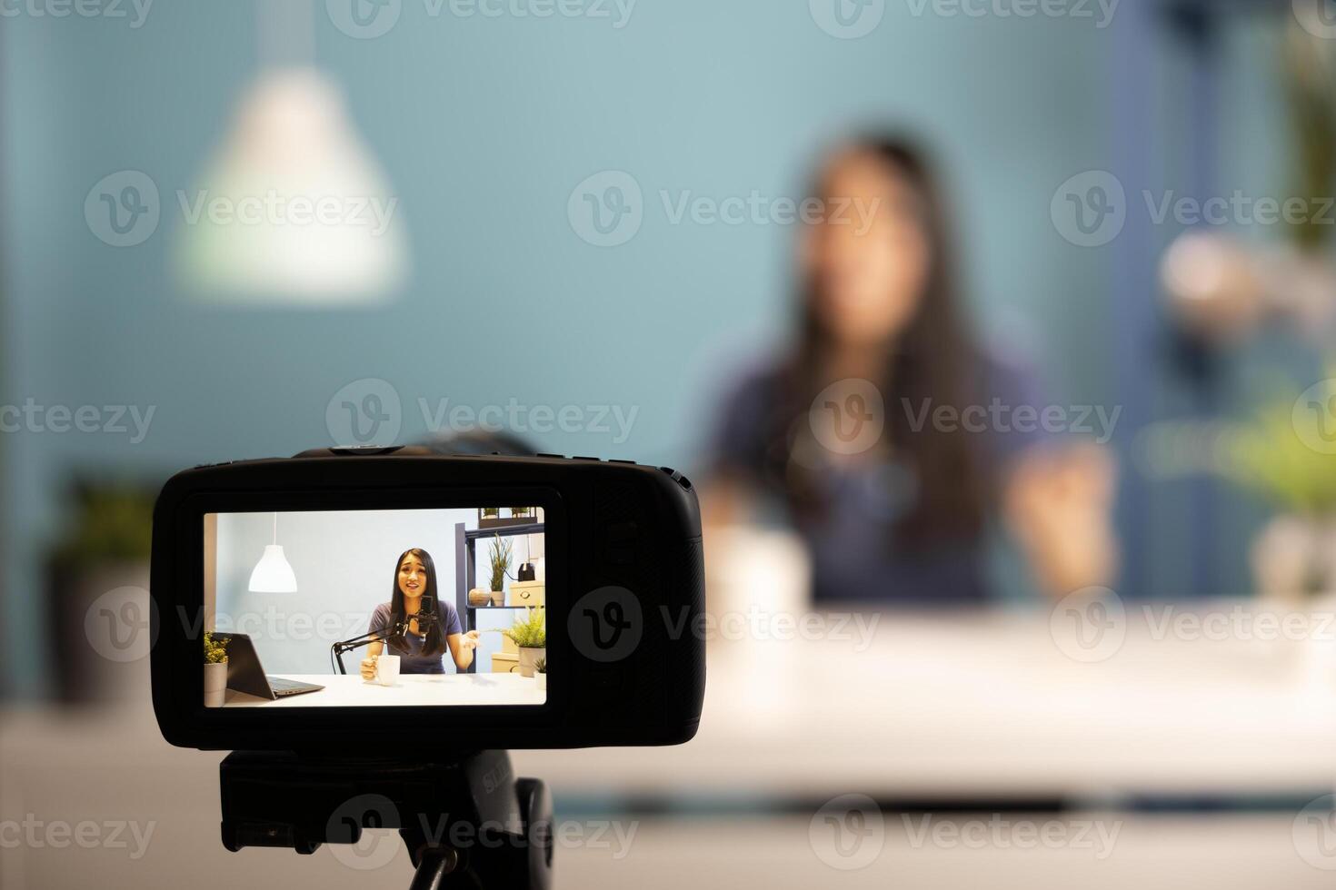 Asian content creator sitting at desk, holding a cup of coffee, speaking into a microphone. Digital camera capturing woman sharing tips on online branding and trends during her live broadcast session. photo