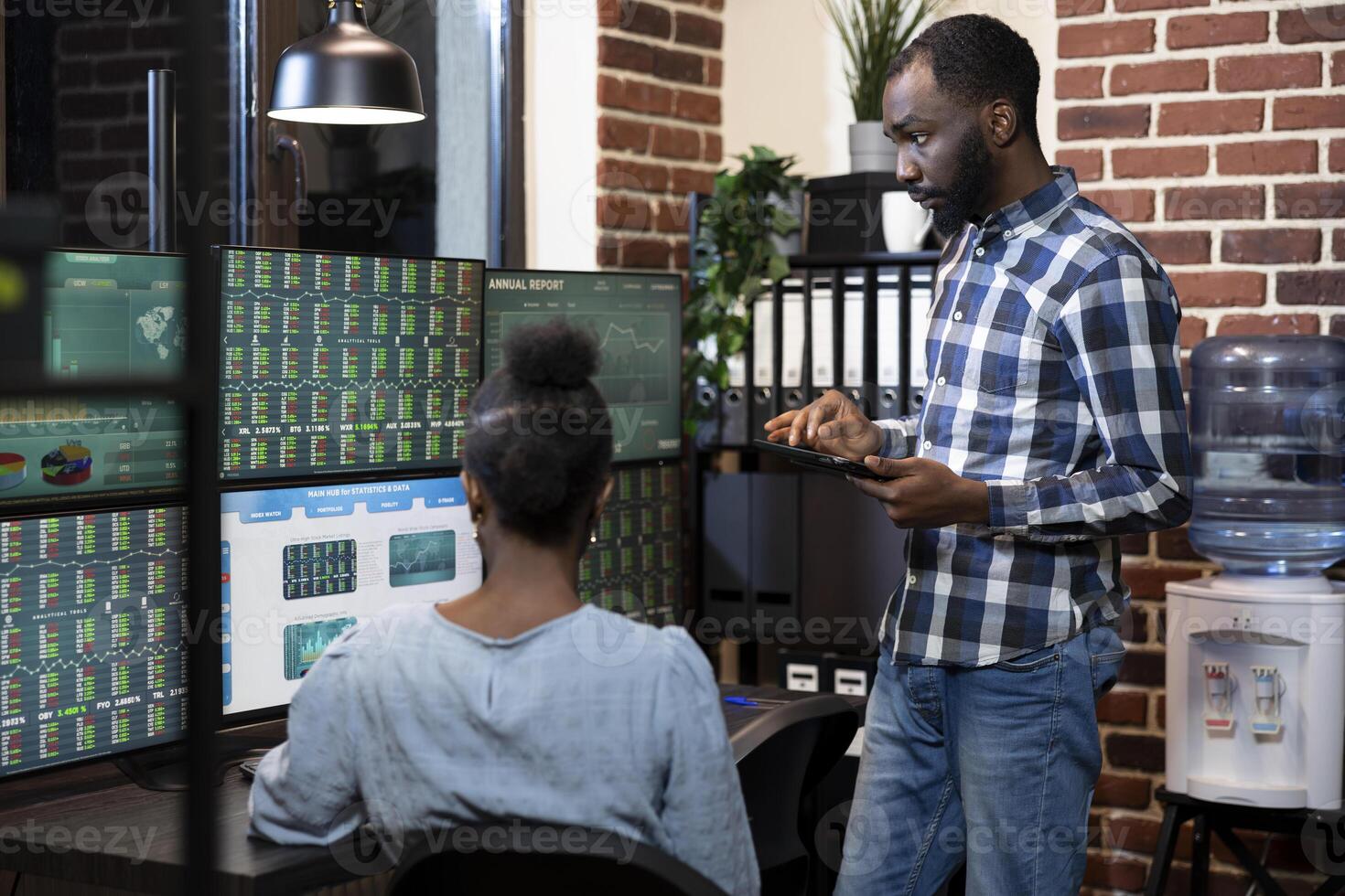 African american team of two stockbrokers working late, reviewing forex charts and financial data on multiple monitors in brick wall office. Male manager with a tablet collaborates with his coworker. photo