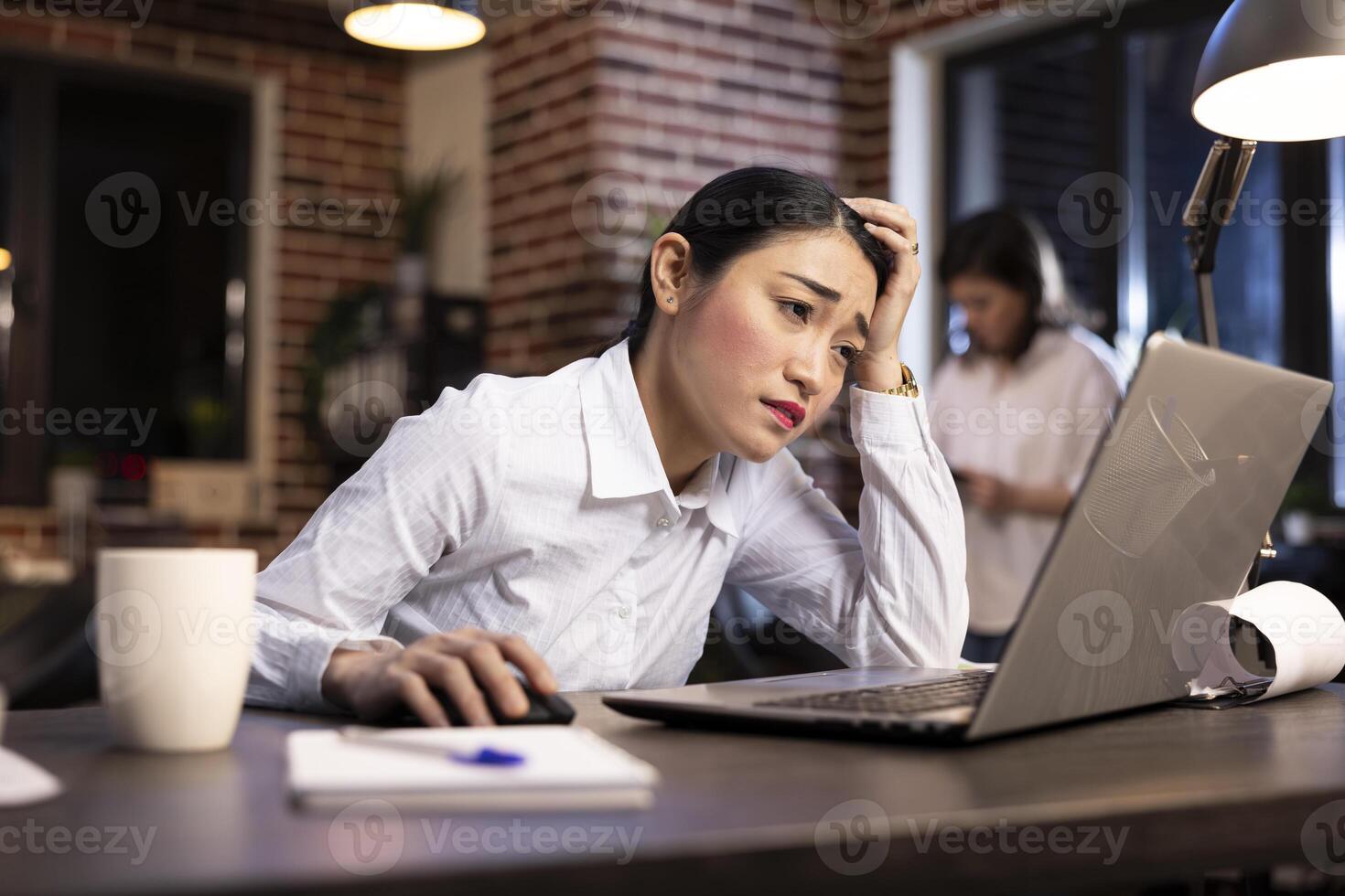 Professional woman working late at desk, exhausted and stressed from busy workday, dealing with pressure of tight deadlines in modern office. Exhausted asian entrepreneur scrolling through her laptop. photo