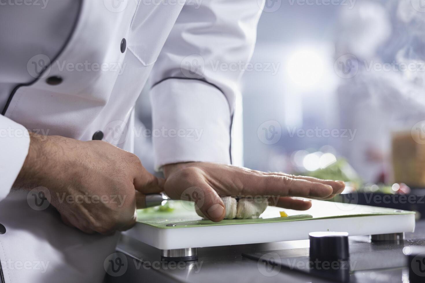 Young chef with hat making crushed garlic with knife on the cutting board, pressing cloves to use condiments for gourmet dish in the restaurant kitchen. Male cook uses spices at counter. Close up. photo