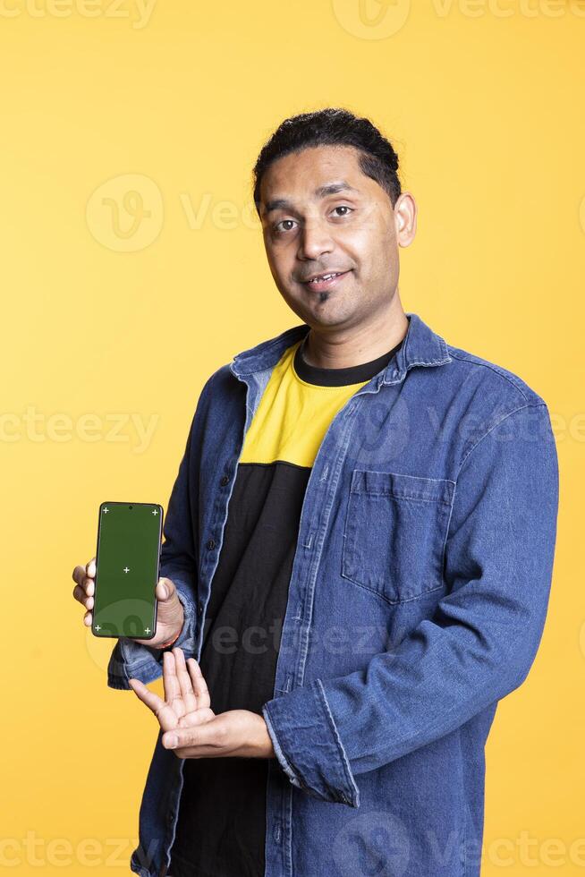 Smiling indian adult presenting mobile phone with isolated mockup screen, staying against studio background. Young casual guy creating a promotion with blank display on gadget. photo