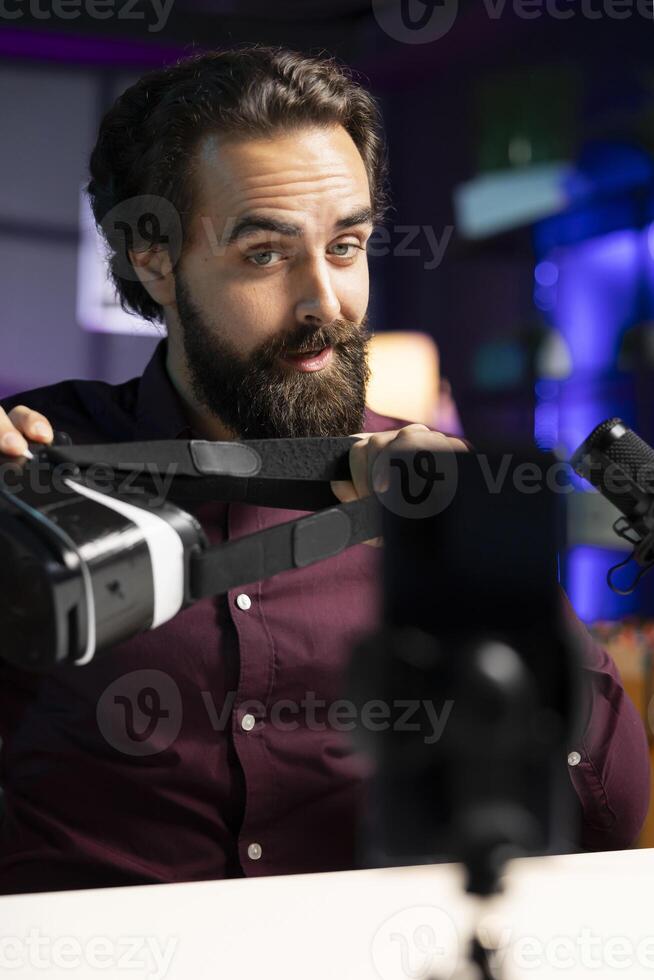 Tech professional reviewing VR glasses in neon lit studio, presenting specifications. Man filming virtual reality headset review for technology fans using cellphone on tripod stabilizer photo