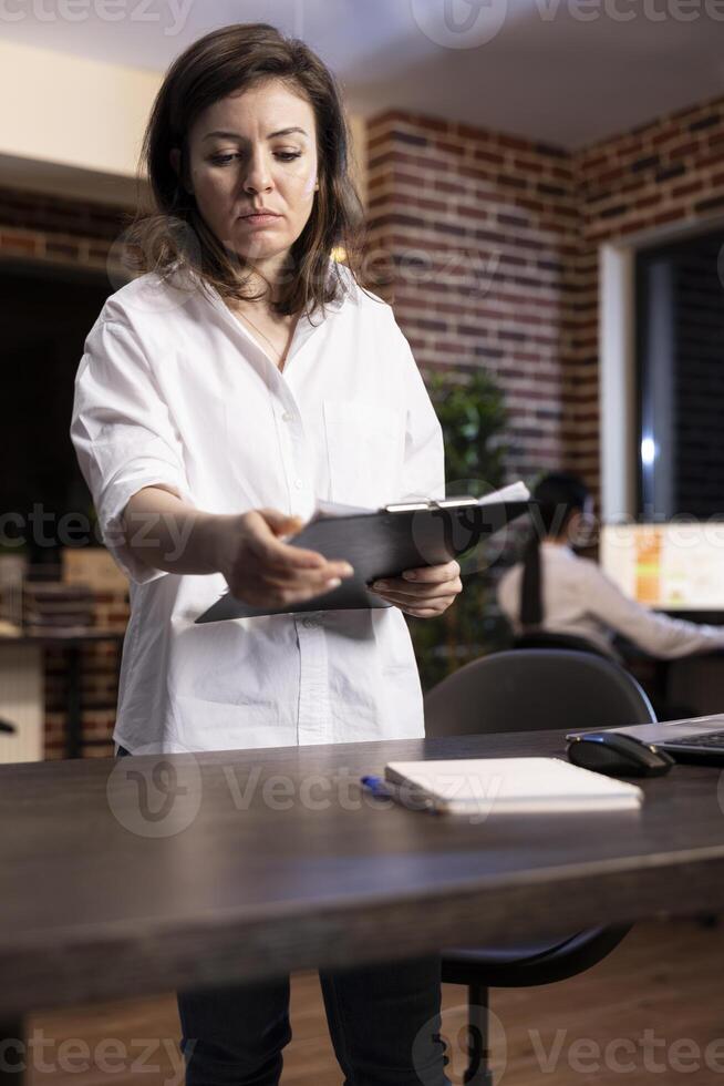 Caucasian female employee stands at her workstation, analyzing documents on a clipboard. She flips through pages, reviewing important information during a late night work session. photo