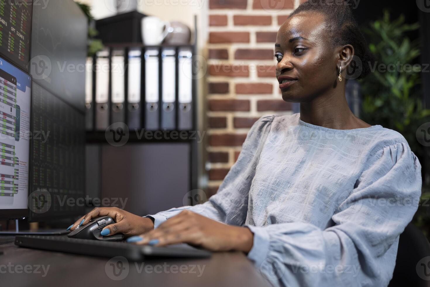 African American trader reviews financial statistics on multiple monitors, studying live stock market data in focused work setting. Black woman at desk, using computer mouse and keyboard in office. photo