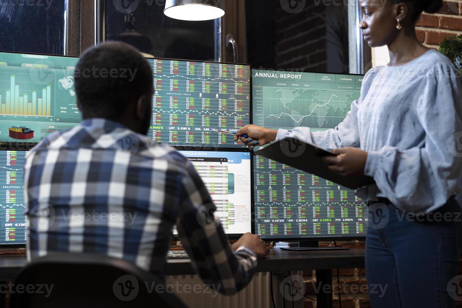 Two traders working late at night in a modern office, collaborating on multiple monitors displaying forex market charts and stock data. African american business partners reviewing stock trading data. photo