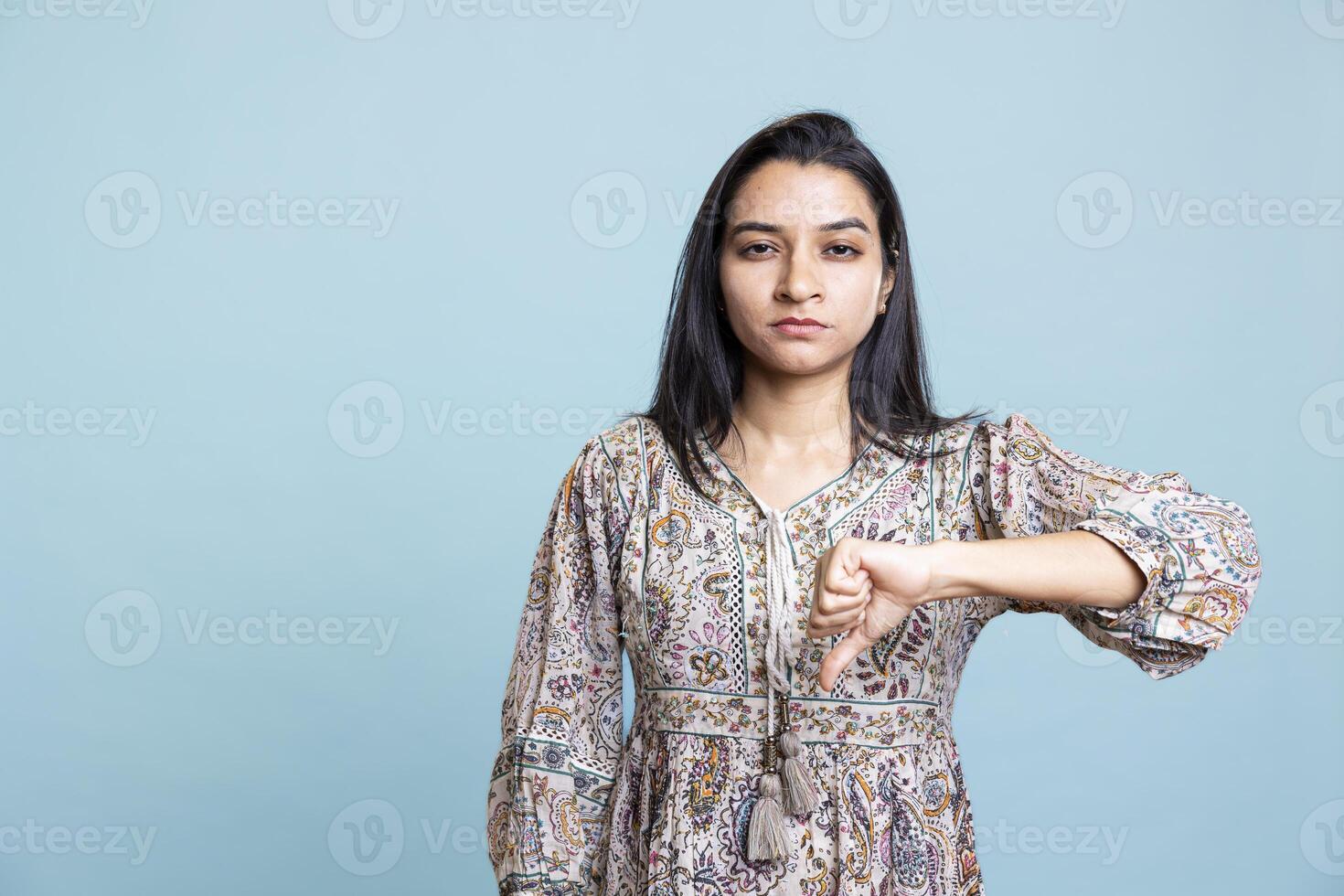 Indian negative model does thumbs down symbol against blue background, expressing disapproval and dissatisfaction. Negative skeptical person gives her bad opinion, being in disagreement. photo