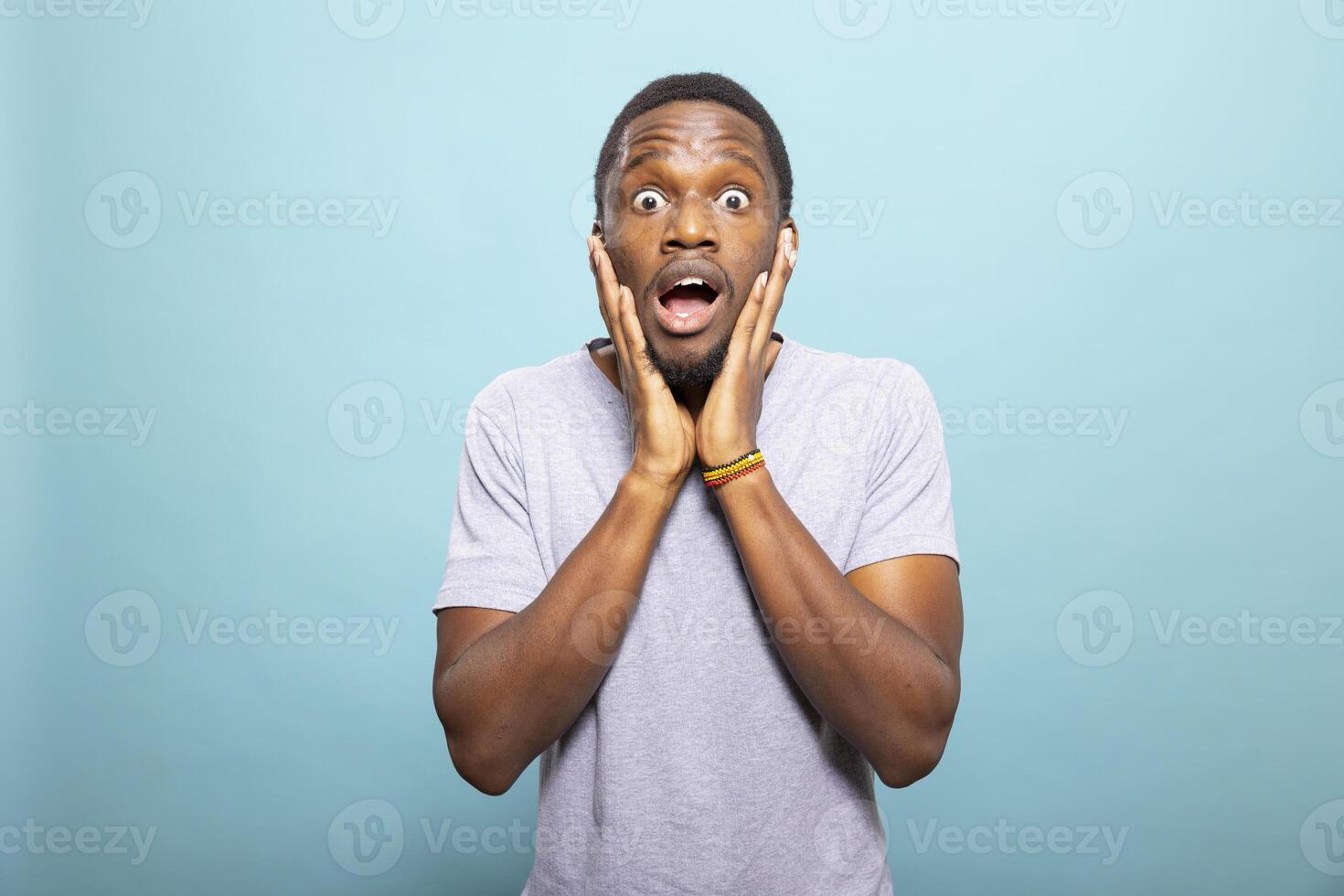 Young african american man with a shocked look, holding his face with two hands in surprise. Black male individual with wide eyes and a startled expression, set against an isolated backdrop. photo