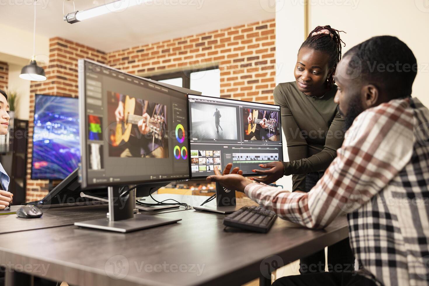 Skilled creative team using multi monitor setup to improve film frame quality in brick wall workspace. Professional male editor working at desk as female teammate assists in enhancing footage visuals. photo