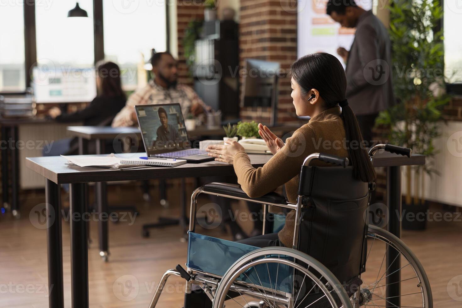Female manager with disability sits in modern office, leading a virtual meeting with a remote colleague. Asian woman in wheelchair uses her laptop to engage in call with african american client. photo