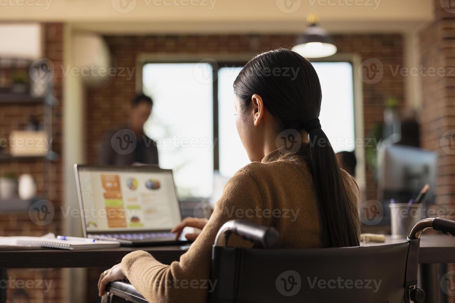 Asian businesswoman with a disability seated at desk with her laptop and notebook, analyzing financial graphs. Female employee in wheelchair catches up with company research work in startup office. photo