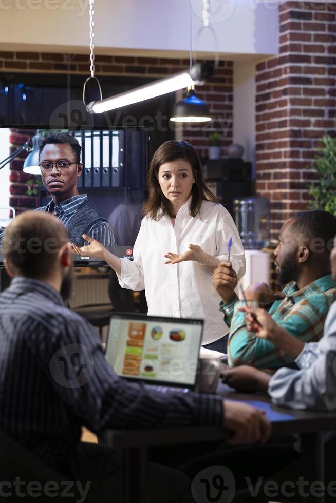 Diverse coworkers in startup office engaged in virtual meeting as caucasian woman leads the discussion. A large monitor displays call with remote male colleague, highlighting modern teamwork. photo
